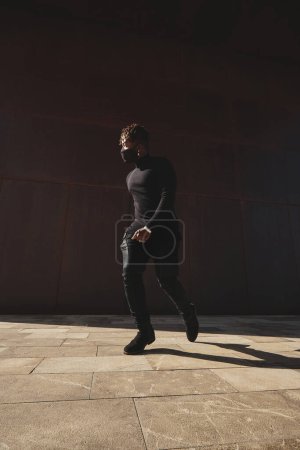 Photo for Side view of anonymous male in black outfit moving on street near wall with shadow - Royalty Free Image