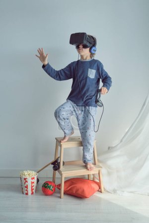 Foto de Cuerpo completo de niño preadolescente positivo en auriculares de realidad virtual y auriculares con joystick de pie en la escalera de taburete cerca de cubo de palomitas de maíz - Imagen libre de derechos