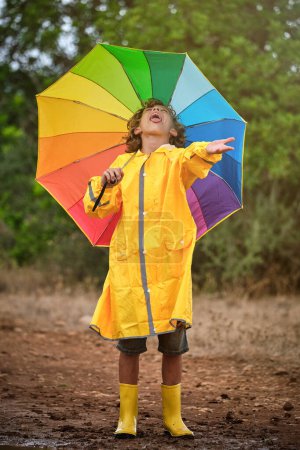 Foto de Cuerpo completo de niño en impermeable amarillo y botas de goma con paraguas colorido disfrutando de la lluvia en el campo - Imagen libre de derechos