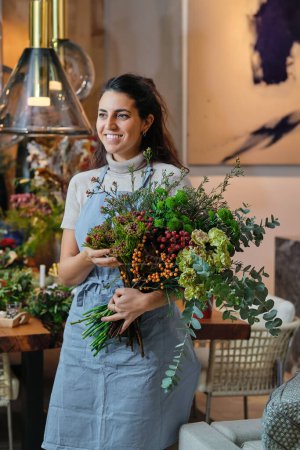Photo for Smiling young woman in blue apron touching arranged flower bouquet and looking away while working in floral shop on blurred background - Royalty Free Image