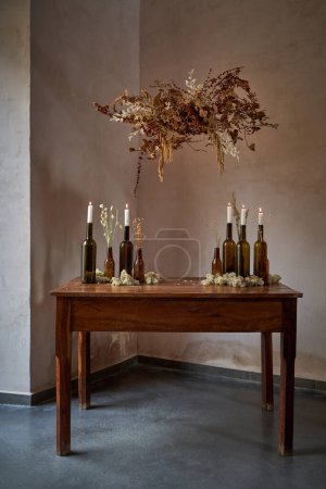 Photo for Retro interior of classic timber table with beer and wine bottles with lit candles and bouquet of dried flowers over - Royalty Free Image
