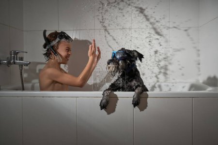 Foto de Vista lateral del chico positivo en la máscara de buceo salpicando agua mientras se divierten en la bañera con perro en gafas de sol en casa - Imagen libre de derechos