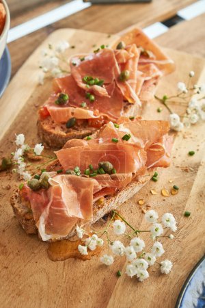 Photo for From above of yummy toasts with appetizing prosciutto and capers and herbs served on wooden cutting board and decorated with delicate white flowers on restaurant terrace - Royalty Free Image