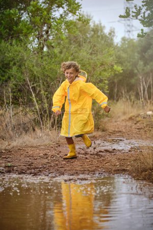 Foto de Niño encantador con impermeable amarillo y botas de goma corriendo al charco sucio y divirtiéndose en el campo - Imagen libre de derechos