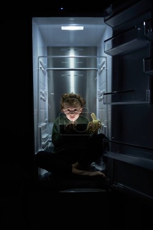 Photo for Full body of child in casual clothes using tablet and eating banana while sitting in fridge with legs crossed in dark kitchen at home - Royalty Free Image