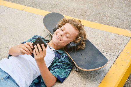 Foto de Desde arriba de niño sonriente en auriculares escuchando música y navegando por teléfono inteligente mientras descansa en el monopatín y tumbado en la acera en la calle - Imagen libre de derechos