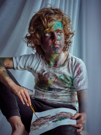 Foto de Retrato de niño creativo en ropa manchada sosteniendo pincel y dibujo sobre papel - Imagen libre de derechos