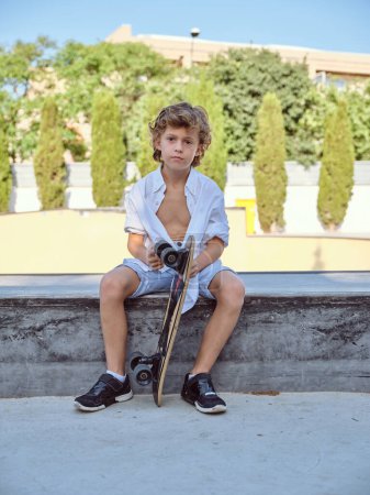 Full body of boy in casual outfit sitting on ramp with skateboard and looking at camera while spending time in park