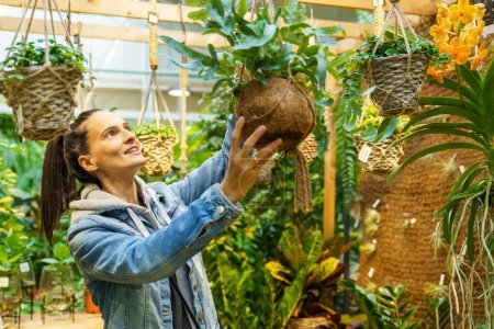 Foto de Feliz cliente femenino hispano mirando hacia arriba mientras que elige las plantas verdes frescas en maceta mientras que elige las flores en la tienda floral espaciosa luz - Imagen libre de derechos