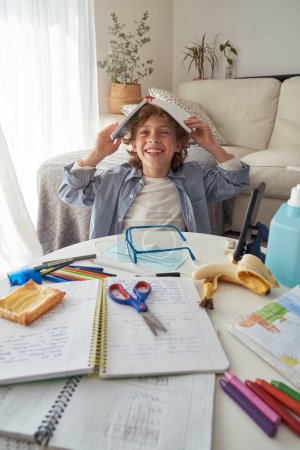Photo for Joyful child with curly hair sitting at table with stationery mask and sanitizer in dispenser and smiling while staying home and doing homework in living room during COVID 19 epidemic - Royalty Free Image