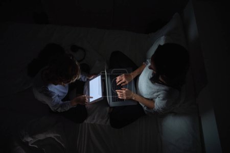 Photo for From above of woman browsing laptop near son surfing tablet while doing homework together in bed in bedroom late at night - Royalty Free Image