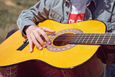 Foto de Desde arriba de la cosecha niño anónimo en ropa casual tocando la guitarra acústica en prado herboso en el día soleado - Imagen libre de derechos