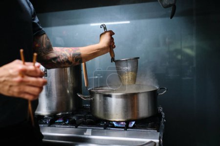 Photo for Side view of crop anonymous tattooed male cook standing near plate and preparing noodles in pot in kitchen - Royalty Free Image