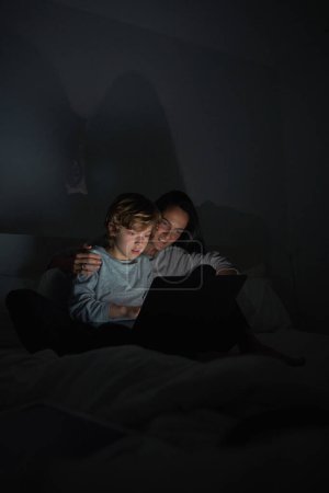 Téléchargez les photos : Femme attentionnée étreignant fils en tenue décontractée tapant sur netbook moderne tout en faisant des devoirs ensemble sur le lit tard dans la nuit - en image libre de droit
