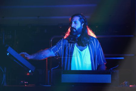 Foto de Profesional masculino en ropa casual tocando canción en la configuración de DJ con mezclador de audio y CDJ mientras realiza música en discoteca oscura - Imagen libre de derechos