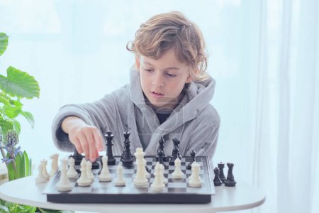 Photo for Concentrated preteen boy in gray terry bathrobe sitting at table and playing chess game in light room - Royalty Free Image