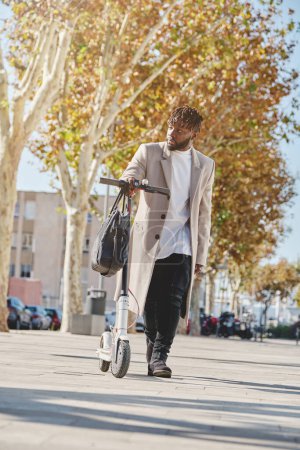 Photo for Ethnic guy in coat looking away while walking with electric scooter on city street in sunlight during fall season - Royalty Free Image