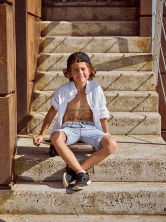 Photo for Full body of smiling preteen skater with curly hair sitting on staircase and looking at camera in summer - Royalty Free Image