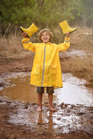 Foto de Niño alegre con impermeable amarillo sosteniendo botas de goma mientras está de pie en el charco y mirando a la cámara durante el tiempo lluvioso - Imagen libre de derechos