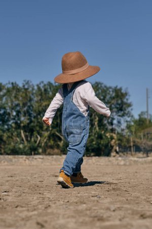 Foto de Vista trasera de un niño divertido irreconocible en denim de moda en general y un gran sombrero caminando en terreno arenoso durante las vacaciones en el campo en un día soleado - Imagen libre de derechos