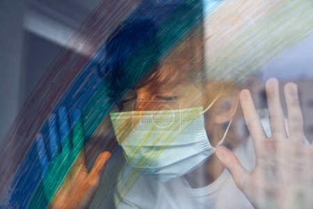 Photo for Preteen kid with curly hair wearing protective face mask standing with closed eyes near window with colorful rainbow during coronavirus epidemic - Royalty Free Image