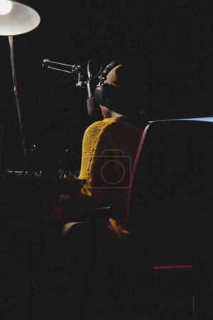 Téléchargez les photos : Vue arrière de l'animateur noir anonyme de radio féminine dans les écouteurs parlant au microphone tout en travaillant dans un studio d'enregistrement moderne sombre - en image libre de droit