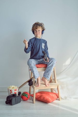 Full body of annoyed kid with blond hair in pajama and headphones playing video game with joystick sitting on wooden stool and clenching fist while losing game