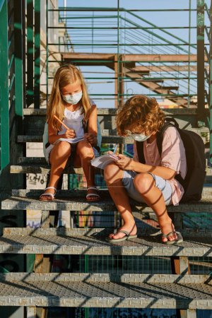 Téléchargez les photos : Pleine longueur de garçon et de fille dans des masques de protection assis sur un escalier en métal et écrivant dans des blocs-notes ensemble pendant l'épidémie COVID 19 - en image libre de droit