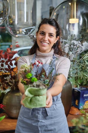 Téléchargez les photos : Joyeux fleuriste féminin en tablier bleu regardant la caméra et démontrant bouquet de fleurs fraîches en fleurs disposées en boîte avec serviette tout en travaillant dans un magasin de fleurs - en image libre de droit