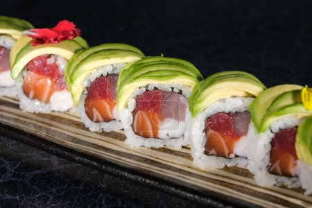 Téléchargez les photos : Grand angle de délicieux rouleaux de sushi uramaki japonais avec des tranches d'avocat sur le dessus servi sur un plateau à la lumière du jour sur une surface noire floue - en image libre de droit