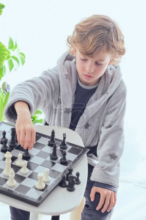 Téléchargez les photos : D'en haut du garçon préadolescent concentré assis à la table et réfléchissant à la stratégie tout en jouant au jeu d'échecs - en image libre de droit