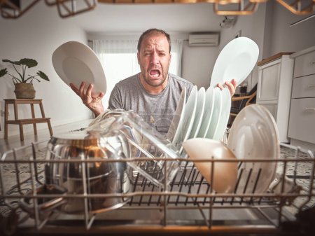 Foto de Hombre infeliz con platos limpios en las manos llorando mientras está de pie cerca de lavavajillas moderno con varios utensilios de cocina durante la rutina del hogar en la cocina - Imagen libre de derechos