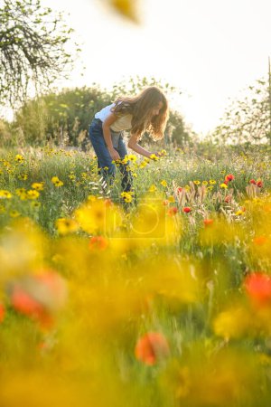 Foto de Chica de cuerpo completo en ropa casual recogiendo flores frescas en prado herboso en el día de verano - Imagen libre de derechos