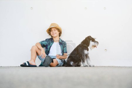 Foto de Retrato de cuerpo completo de niño positivo en ropa casual y sombrero sentado cerca adorable perro miniatura Schnauzer contra la pared en la ciudad - Imagen libre de derechos