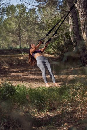Photo for Female athlete exercising leaning back holding on to TRX bands on glade in woods near grass and trees - Royalty Free Image