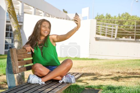 Téléchargez les photos : Souriant jeune femme en vêtements décontractés s'amuser et prendre l'autoportrait au téléphone tout en étant assis sur un banc en bois dans le parc de la ville le jour lumineux d'été - en image libre de droit