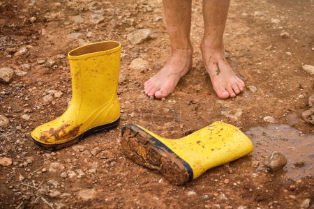 Téléchargez les photos : Enfant méconnaissable avec les jambes couvertes de boue debout près de bottes de gomme jaune sale sur le chemin dans les bois le jour de l'automne pluvieux - en image libre de droit
