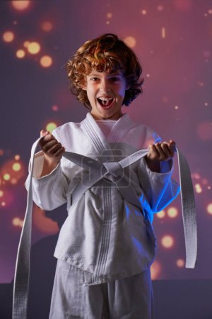 Téléchargez les photos : Enfant joyeux en kimono blanc serrant la ceinture et regardant la caméra tout en restant debout en studio avec des lumières lumineuses pendant l'entraînement du judo - en image libre de droit
