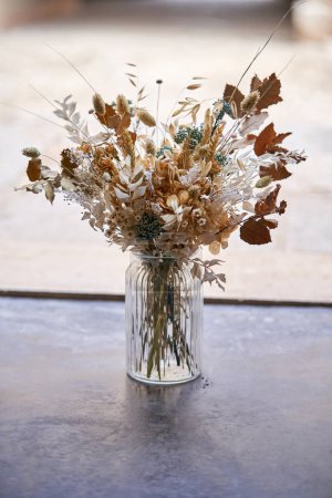 Foto de Ramo de flores secas con ramitas y hojas en jarrón de vidrio colocado en la mesa en la terraza del edificio en el día de verano - Imagen libre de derechos