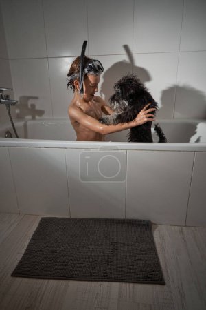 Téléchargez les photos : Vue latérale du garçon en masque de plongée jouant avec le chien noir assis dans la baignoire pendant la routine quotidienne dans la salle de bain - en image libre de droit