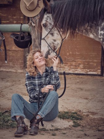 Foto de Cuerpo completo de hembra encantada acariciando caballo manchado blanco con brida mientras está sentado cerca de establo de madera en el área rural - Imagen libre de derechos