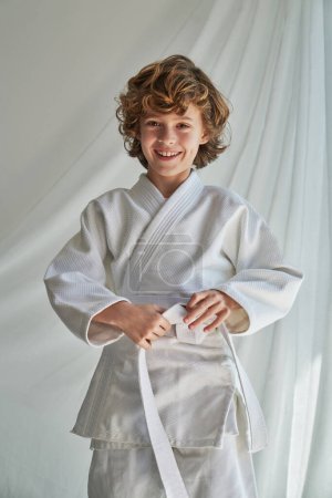Photo for Smiling curly haired boy in white kimono standing near curtain and tying belt while looking at camera before judo training - Royalty Free Image