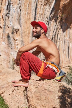 Photo for Side view of muscular shirtless man wearing climbing harness chalk bag red pants and cap looking away and sitting near high mountain on sunny day - Royalty Free Image