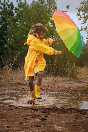 Foto de Cuerpo completo de niño excitado en impermeable amarillo y botas de goma que se divierten mientras corre a través de charcos en el bosque en el día lluvioso - Imagen libre de derechos