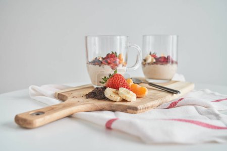 Foto de Composición de yogur fresco con chocolate rallado y mandarina de plátano en rodajas y fresa colocados en la tabla de cortar en la servilleta blanca - Imagen libre de derechos