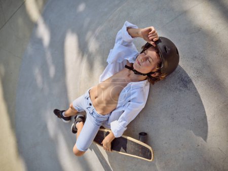 Von oben ein kleiner Junge in Freizeitkleidung und Helm auf Asphalt liegend und mit dem Skateboard in der Hand in den Himmel blickend