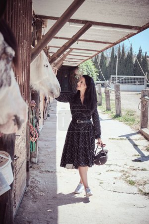 Foto de Cuerpo completo de contenido ecuestre con casco acariciando caballo blanco de pie en el recinto en el día soleado de verano en el área rural - Imagen libre de derechos
