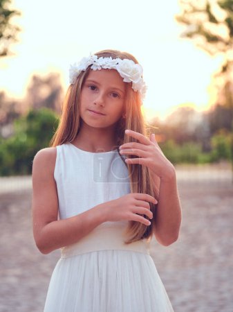 Photo for Peaceful preteen girl in floral wreath and white dress gently touching long hair and looking at camera while standing on background of sunset sky in park - Royalty Free Image