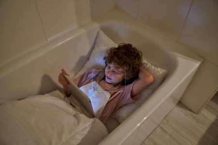 Photo for From above of child resting in bathtub with pillow and blanket while watching video on tablet in bathroom at home - Royalty Free Image