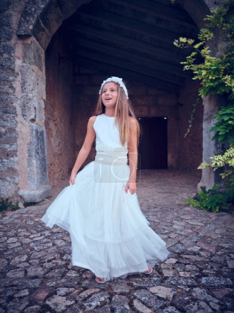 Photo for Full length of pretty teenager wearing stylish white maxi gown and floral diadem standing in front of aged stone castle entrance and looking aside - Royalty Free Image